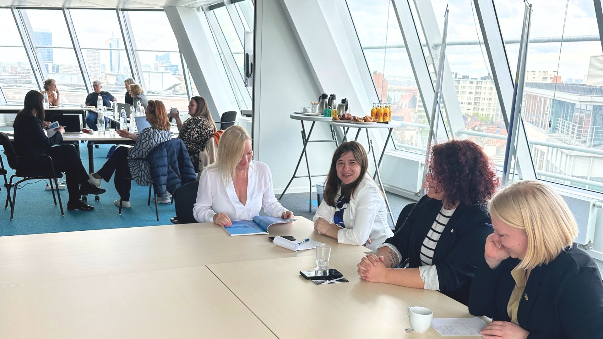 A selection of women sat around desks, in a room with windows looking out over a city scape.