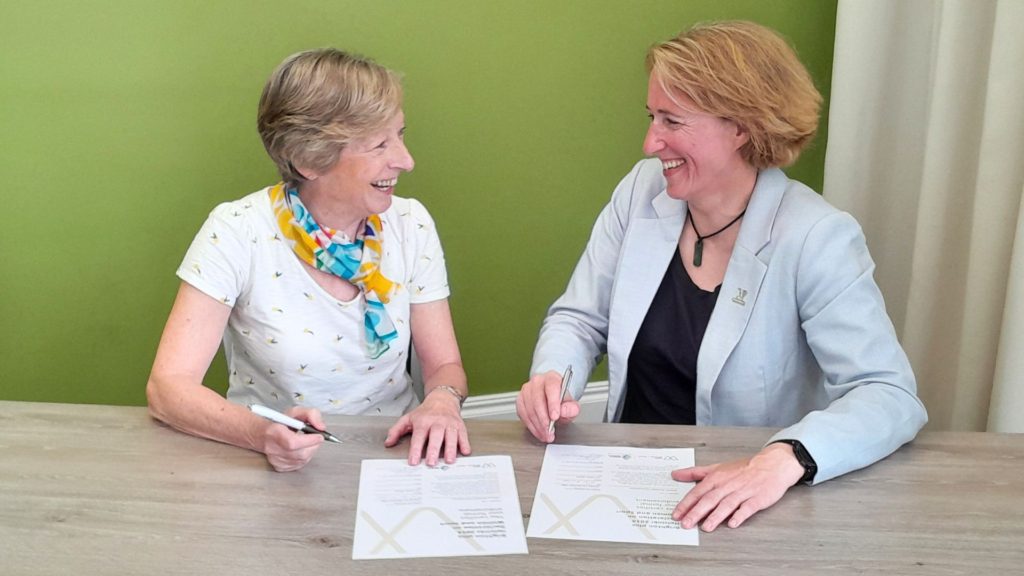 Lisa O'Keefe MBE and Dame Liz Nicholl DBE smile at each other while signing the Brighton Plus Helsinki Declaration