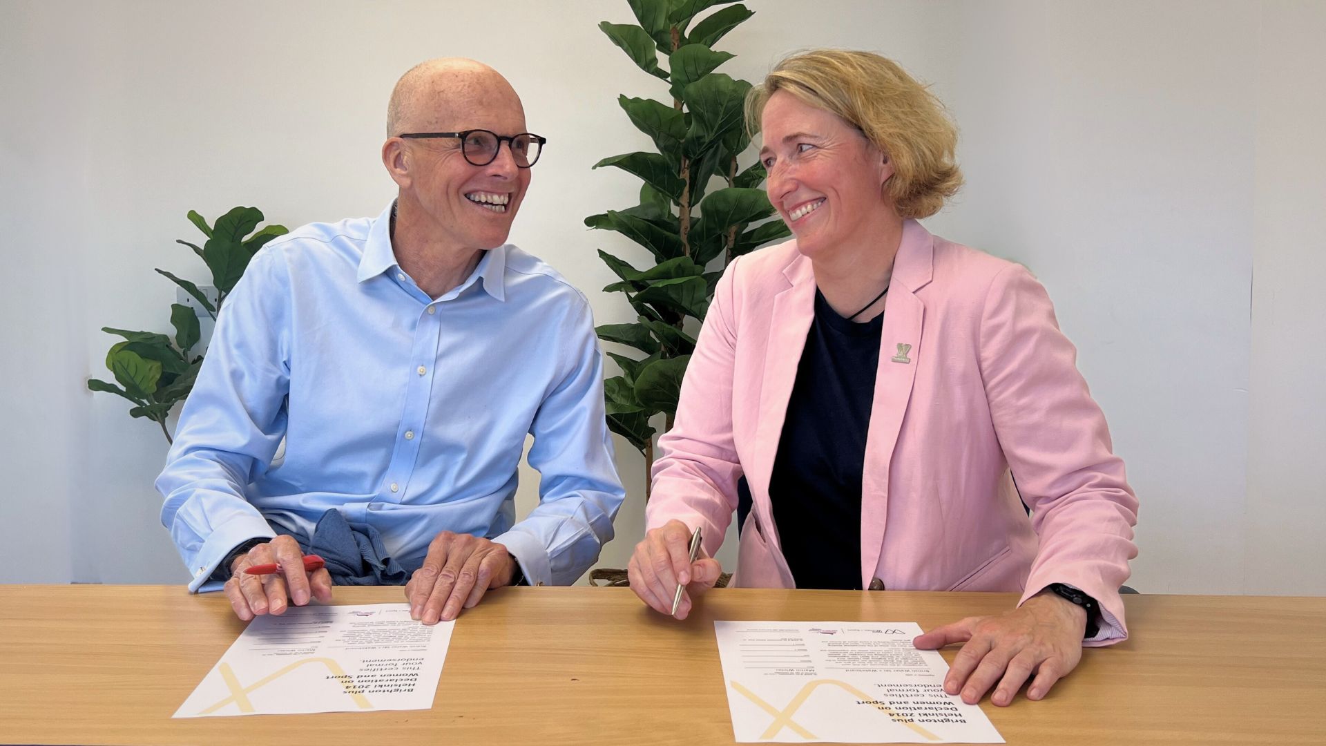 Martin Winter, Chair of British Water Ski and Wakeboard, sits next to Lisa O'Keefe, Secretary General of IWG Woman and Sport. They are both smiling at the camera as they have just signed the Brighton Plus Helsinki Declaration.